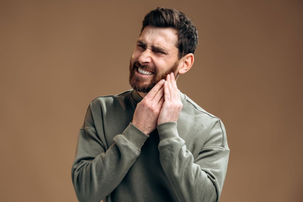 Man in a green sweatshirt clutches his jaw with a pained, grimacing expression, eyes closed.