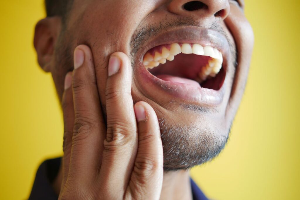 Close-up of a man pressing his cheek with his fingers, mouth open to reveal teeth against a yellow background.