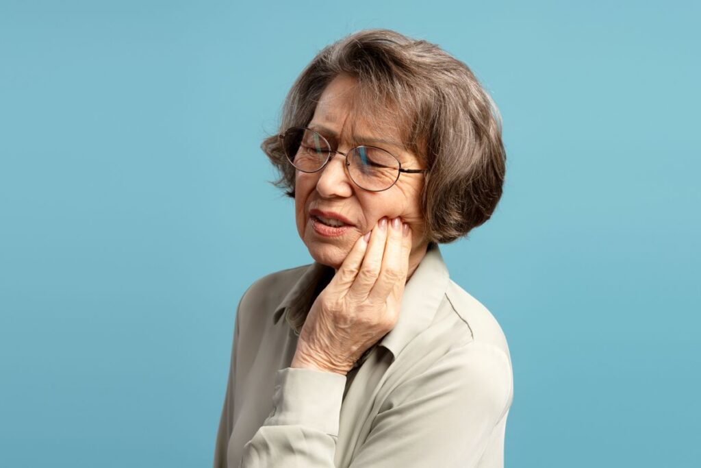 Older woman with short gray hair and glasses pressing her cheek in pain against a blue background.