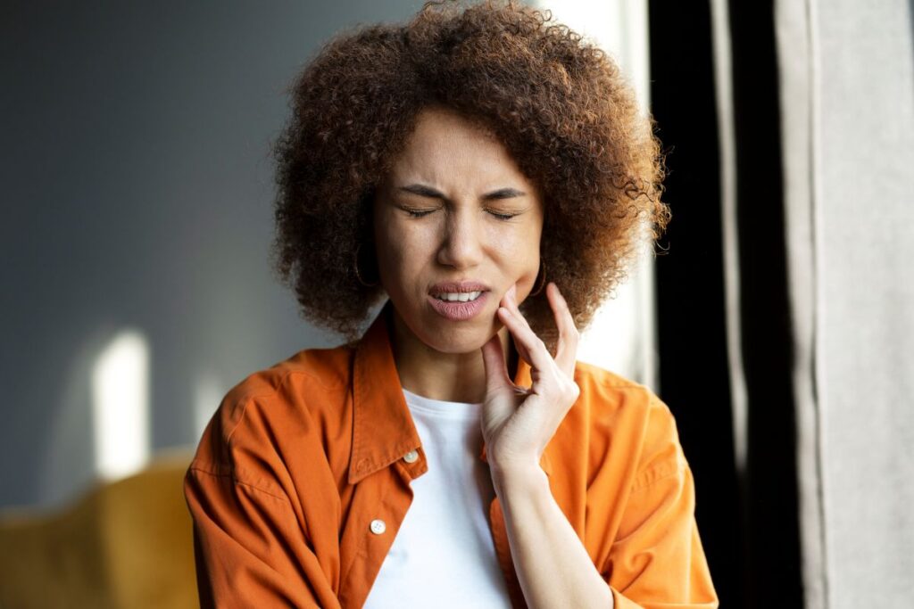Woman with curly hair wearing an orange shirt touching her jaw in apparent toothache while seated by a window