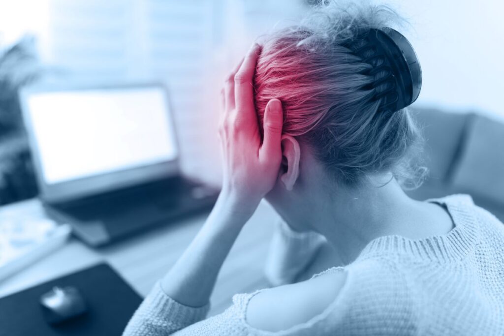 Person wearing headphones holds their head in frustration at a desk with a laptop nearby.