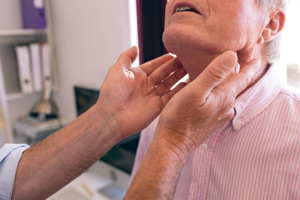 Clinician palpating an elderly man's neck during a medical throat examination in a clinic setting.