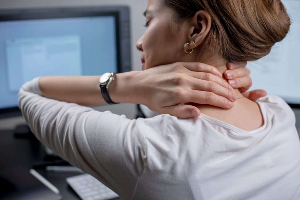 Woman seated at a desk massages the back of her neck with both hands, indicating neck stiffness or pain near a computer monitor in the background.
