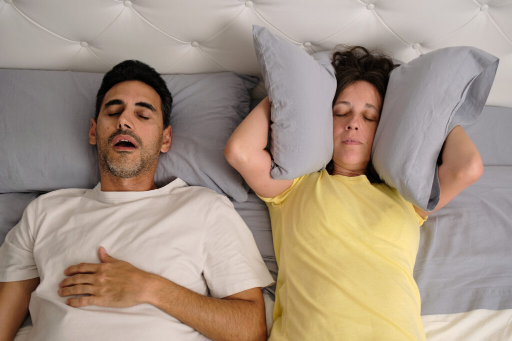 Woman covering her ears with a pillow while her partner snores with his mouth open in bed